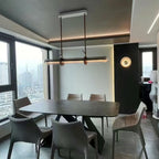 A Clérachic glass tube pendant hung from a white ceiling plate above a dark stone dining table, with grey chairs and a city view outside.