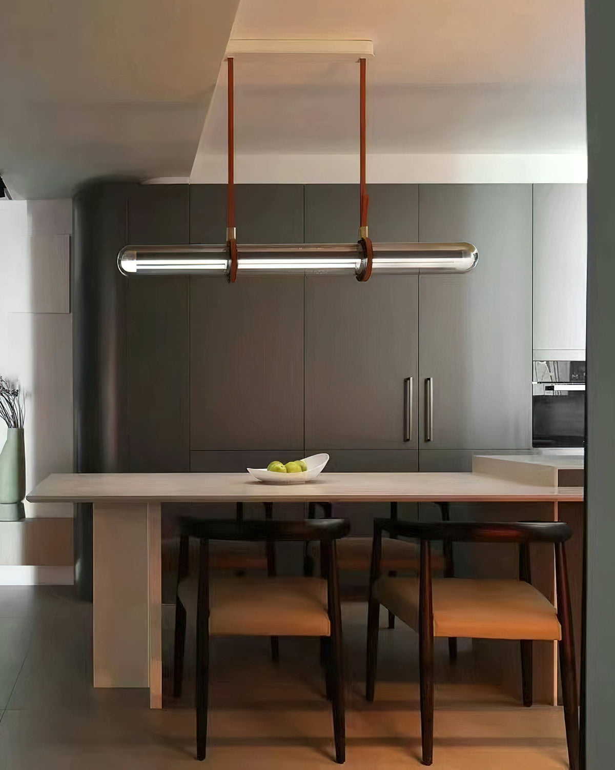 A Clérachic glass tube pendant hung from straps above a pale table with a bowl of limes, in front of dark grey cabinets.