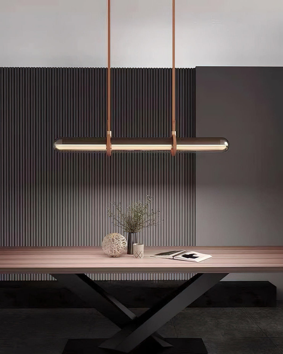 A Clérachic glass tube pendant lit above a pale wooden table with a vase and a book, against a dark ribbed wall.