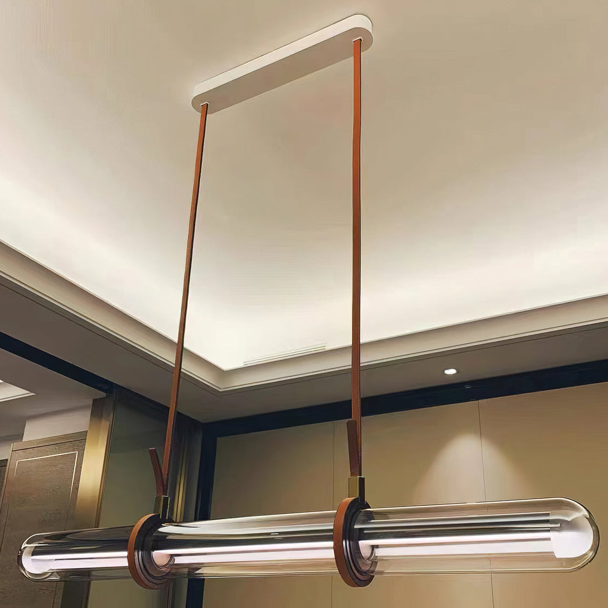 Looking up at a Clérachic glass tube pendant, with two long brown leather straps running to a white plate on a ceiling with a lit edge.