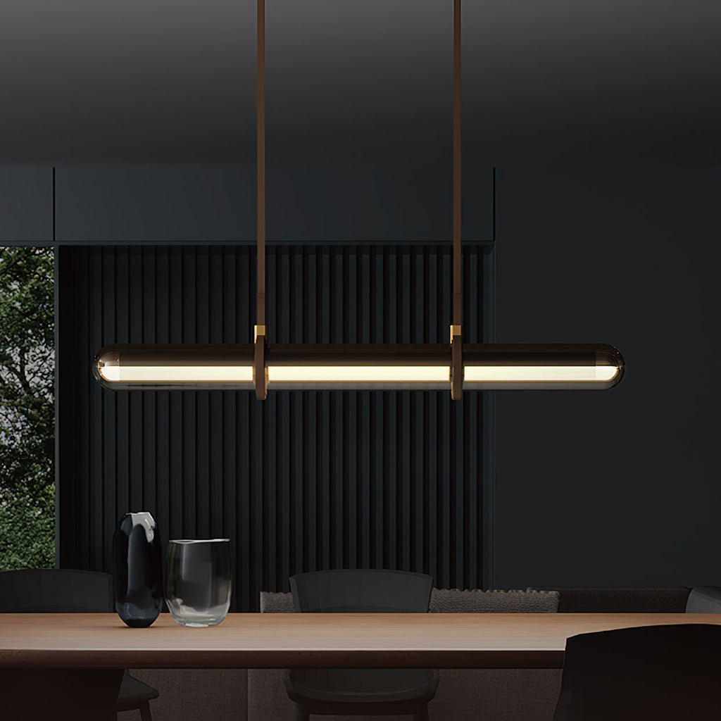 A Clérachic glass tube pendant glowing warm above a wooden table in a very dark room, with a glass and a vase on the table.