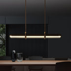 A Clérachic glass tube pendant glowing warm above a wooden table in a very dark room, with a glass and a vase on the table.