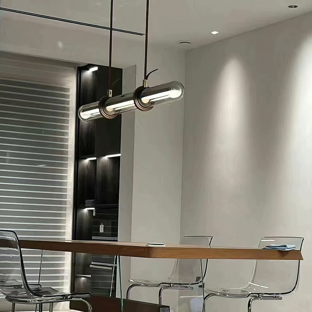 A Clérachic glass tube pendant lit above a long wooden table with clear acrylic chairs, beside a dark shelving unit.