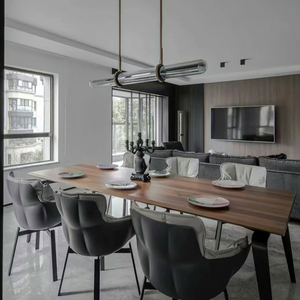 A Clérachic glass tube pendant seen at an angle above a wooden dining table, with grey quilted chairs and a sofa in the room behind.