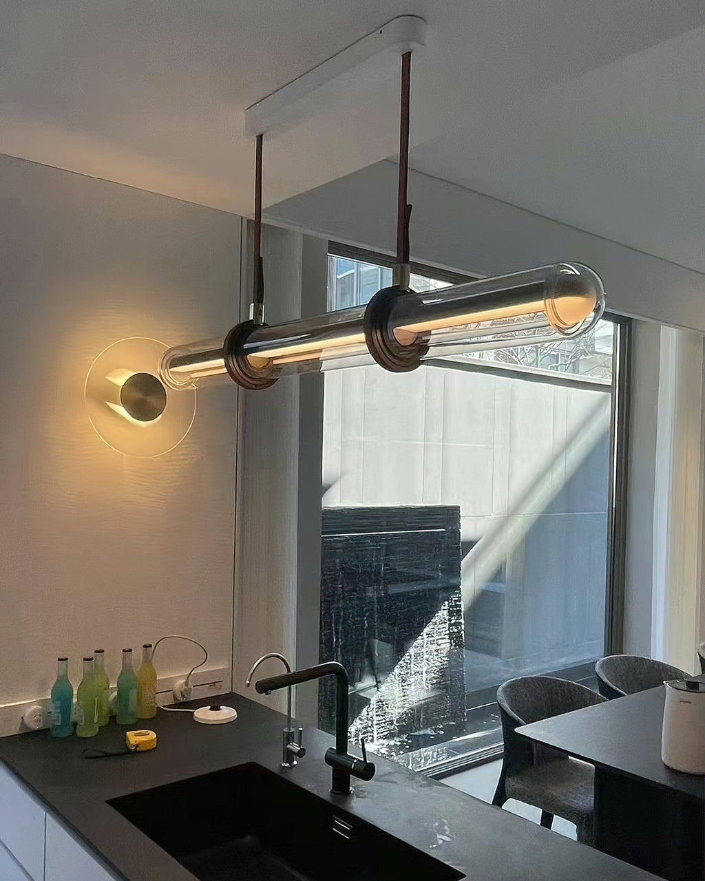A Clérachic glass tube pendant in a kitchen, hung from a white ceiling plate by a window and a sink, with a round wall light beside it.