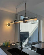 A Clérachic glass tube pendant in a kitchen, hung from a white ceiling plate by a window and a sink, with a round wall light beside it.