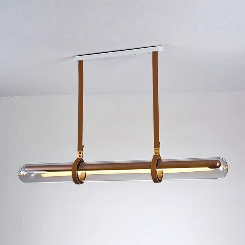 A Clérachic glass tube pendant on tan leather straps from a white ceiling plate, glowing warm against a plain white wall.