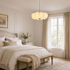 Clérachic glass-disc pendant hanging over a bedroom with a tufted bench and a cream bed, beside a window with linen curtains.