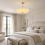 Brass-stemmed Clérachic pendant in a bedroom with French doors, a cream bed and a pale upholstered bench at its foot.