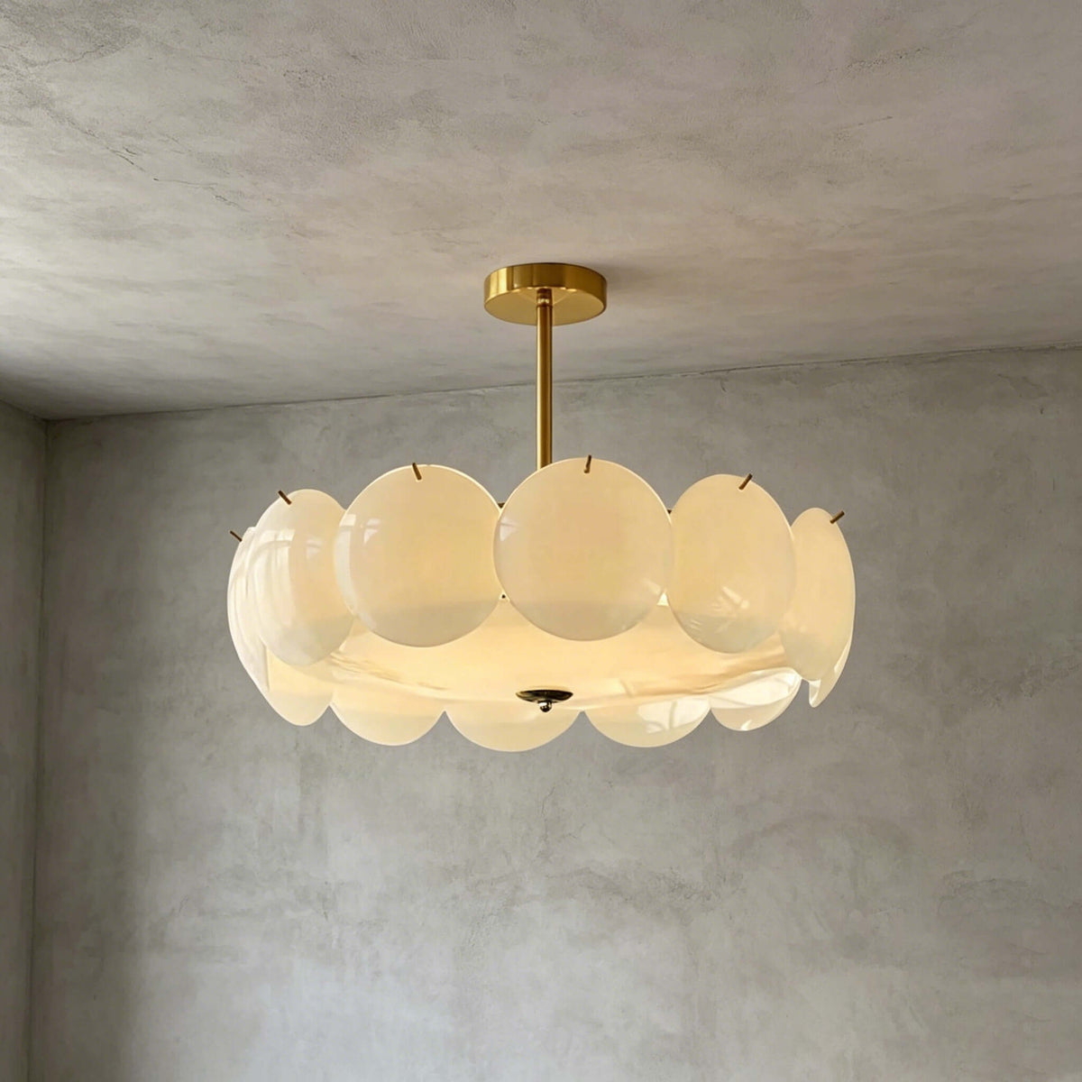 Larger brass-finish Clérachic pendant with a wide ring of white glass discs, lit softly against grey plaster.