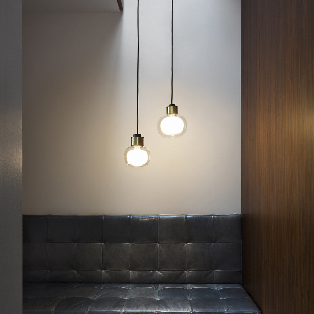 Two small Clérachic glass pendants with gold collars glow above a tufted dark leather banquette and a walnut panel.