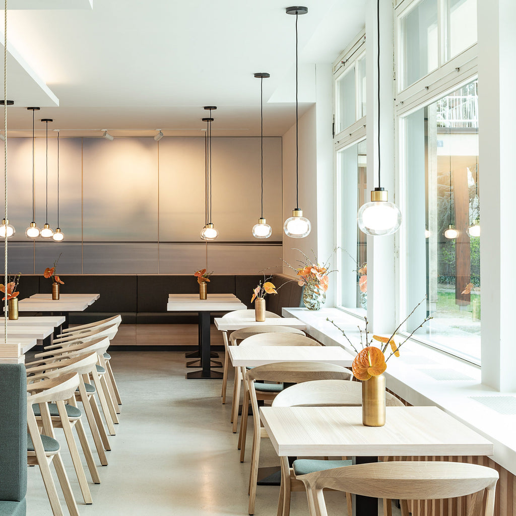 Clear-glass pendants from Clérachic hang at different heights beside tall windows in a bright café with pale wooden chairs.