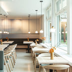 Clear-glass pendants from Clérachic hang at different heights beside tall windows in a bright café with pale wooden chairs.