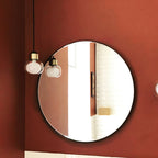 Two Clérachic glass pendants with gold collars hang beside a round black-framed mirror on a terracotta-red wall.