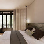 A single Clérachic glass pendant hangs near a tall window in a bedroom, over the foot of a white bed with a grey throw.