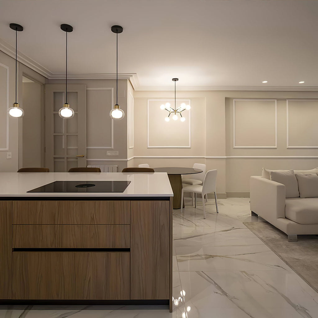 Three Clérachic glass pendants hang over a kitchen island with a wood-fronted base, beside a white marble floor and dining table.