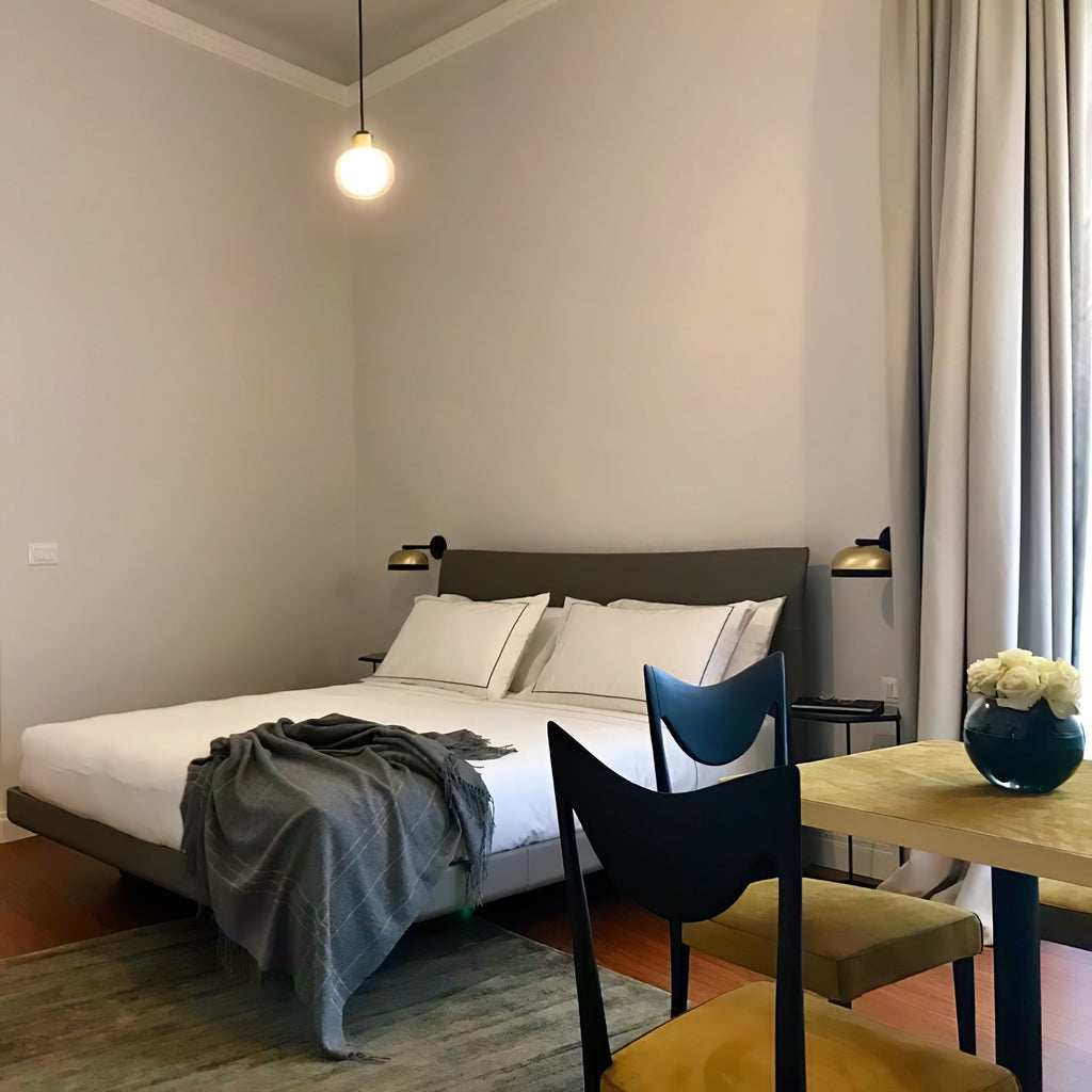 A single Clérachic glass pendant glows above a bed with a grey throw, beside a wooden dining table in the same room.