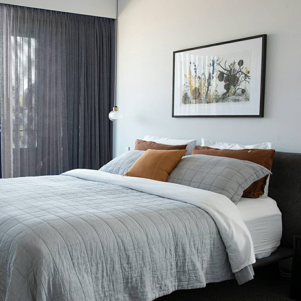 A Clérachic glass pendant hangs beside a bed with grey quilted bedding and rust cushions, below a framed botanical print.