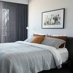 A Clérachic glass pendant hangs beside a bed with grey quilted bedding and rust cushions, below a framed botanical print.
