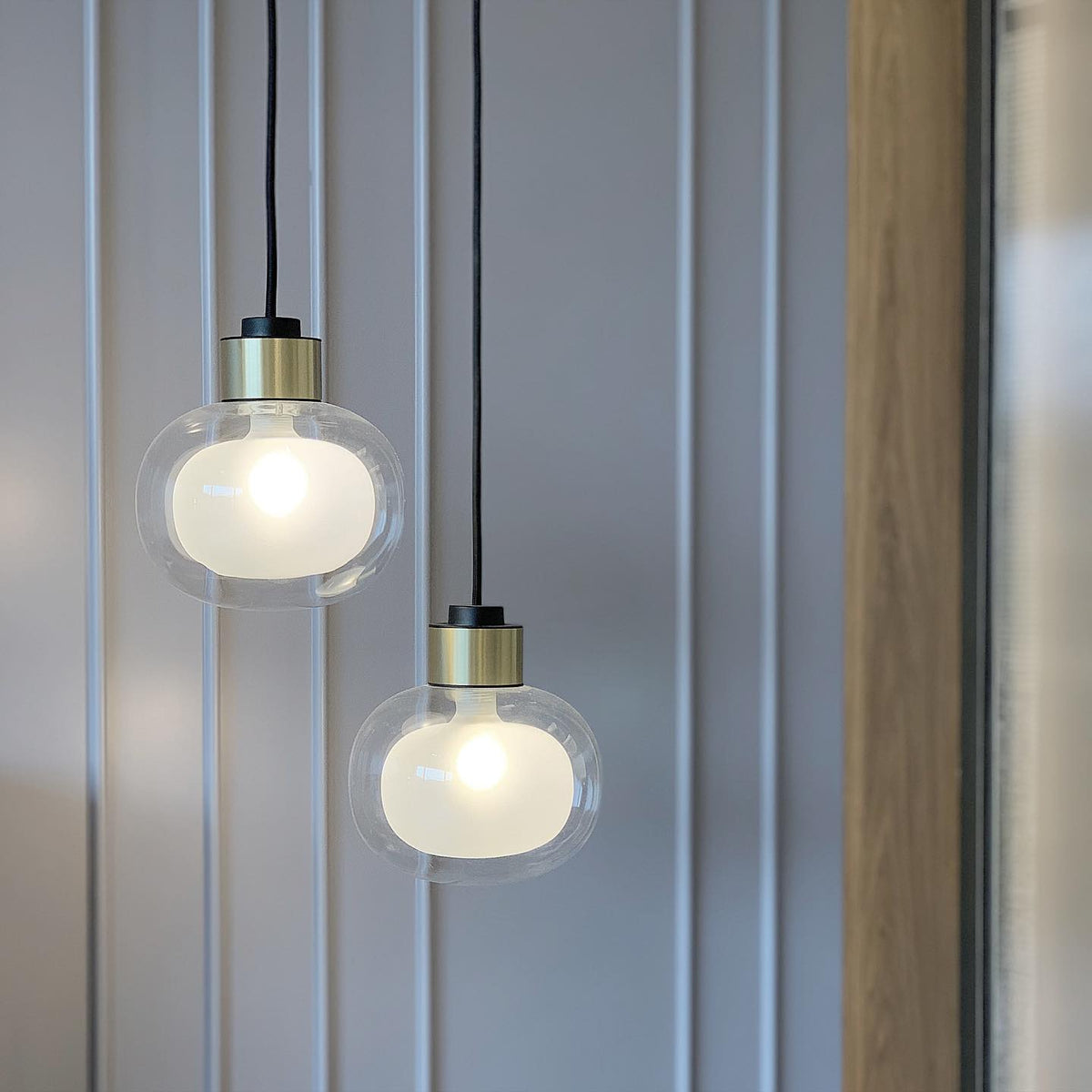 Close view of two Clérachic glass pendants with gold collars and a frosted inner glow, hung against a pale panelled wall.