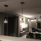 A Clérachic glass pendant hangs in the foreground of a dim living and dining room, with softly lit curtains and a stone wall behind.