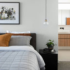 A single Clérachic glass pendant hangs by a bedside with grey quilted bedding, rust cushions and a framed botanical print behind.