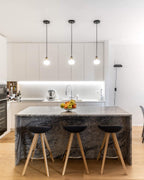 Three Clérachic glass pendants hang in a row over a stone-fronted kitchen island with black wooden-legged stools.
