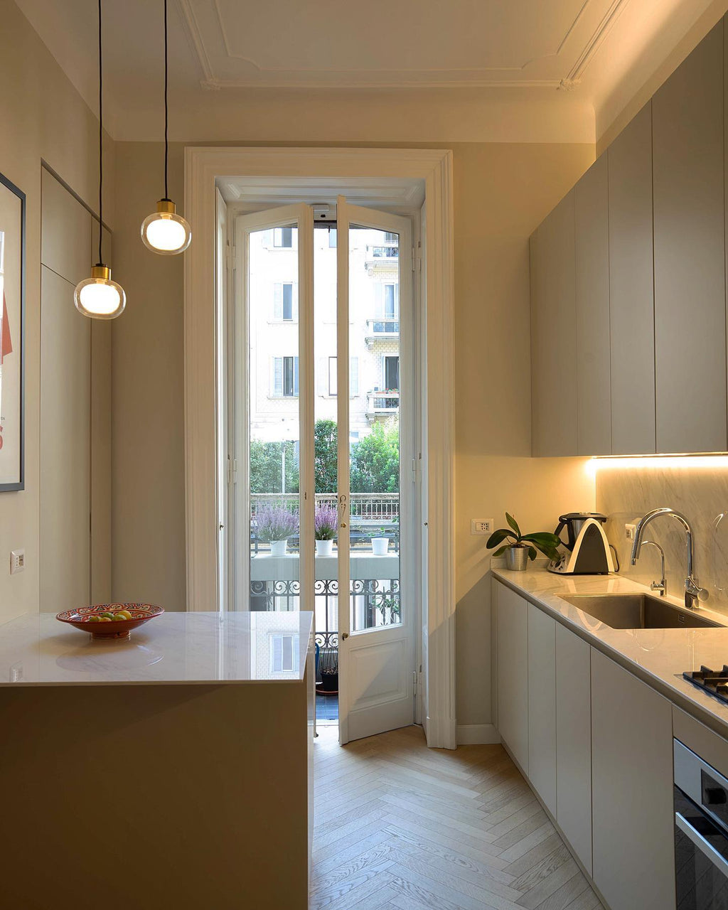 Two Clérachic glass pendants hang in a kitchen with tall French windows and herringbone parquet flooring.