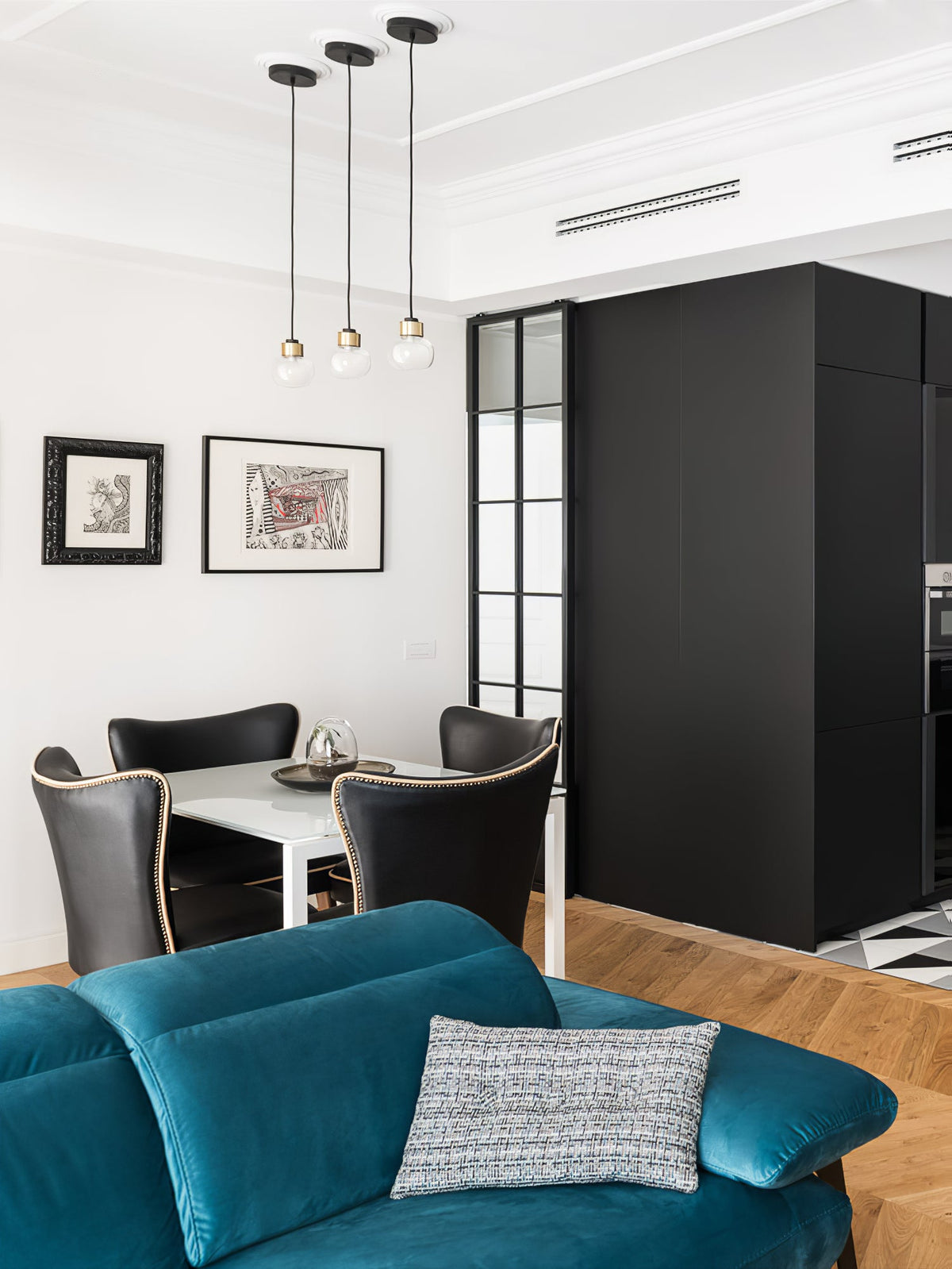 Three Clérachic glass pendants hang over a dining table in a white room with framed prints, a black cabinet and a teal velvet sofa.