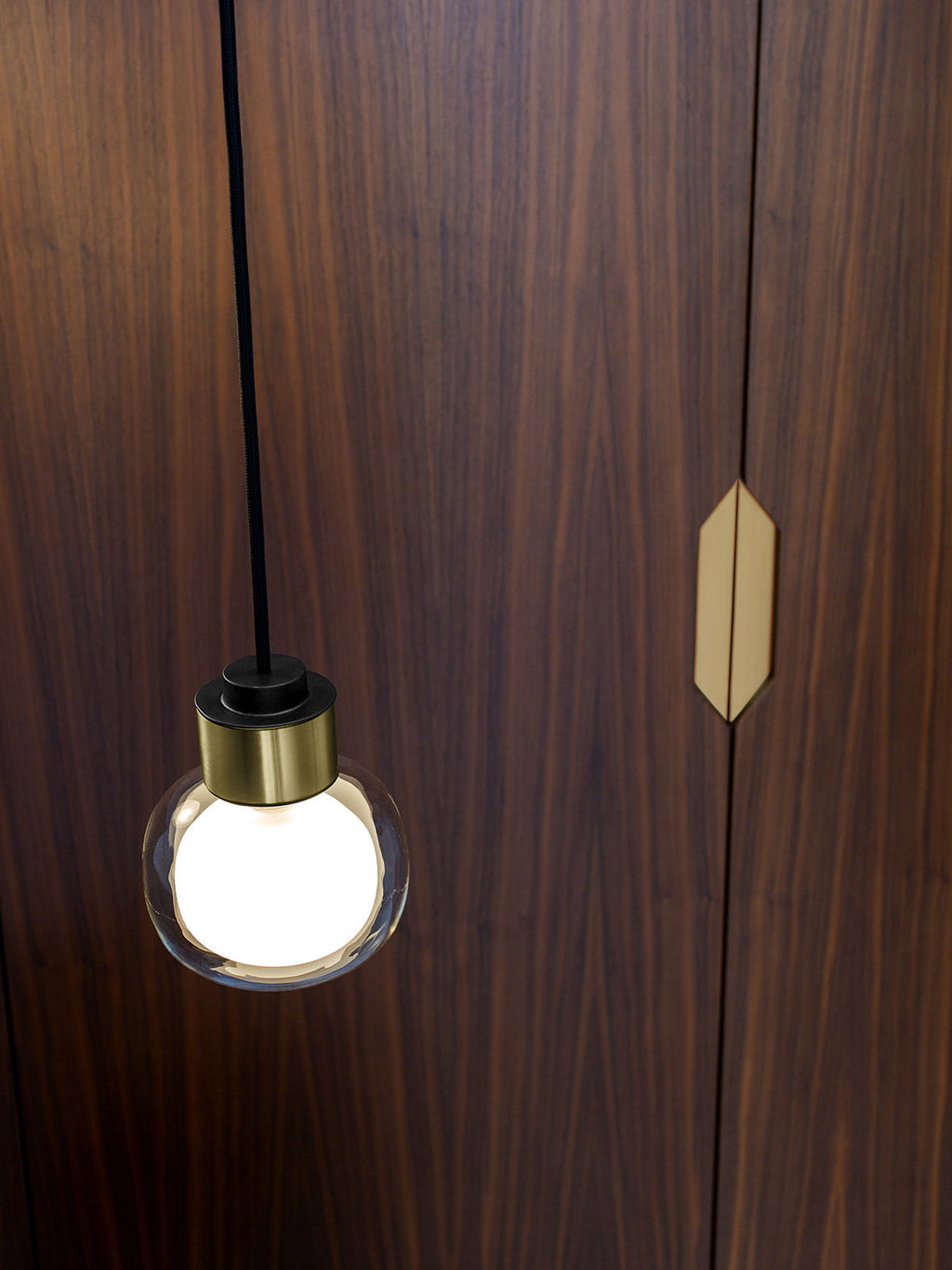 Close view of a Clérachic glass pendant with a gold collar and a frosted globe, against a walnut door with a brass handle.