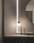 A Clérachic glass pendant glows beside a pale round mirror above a textured stone vanity with a diffuser and a soap bottle.
