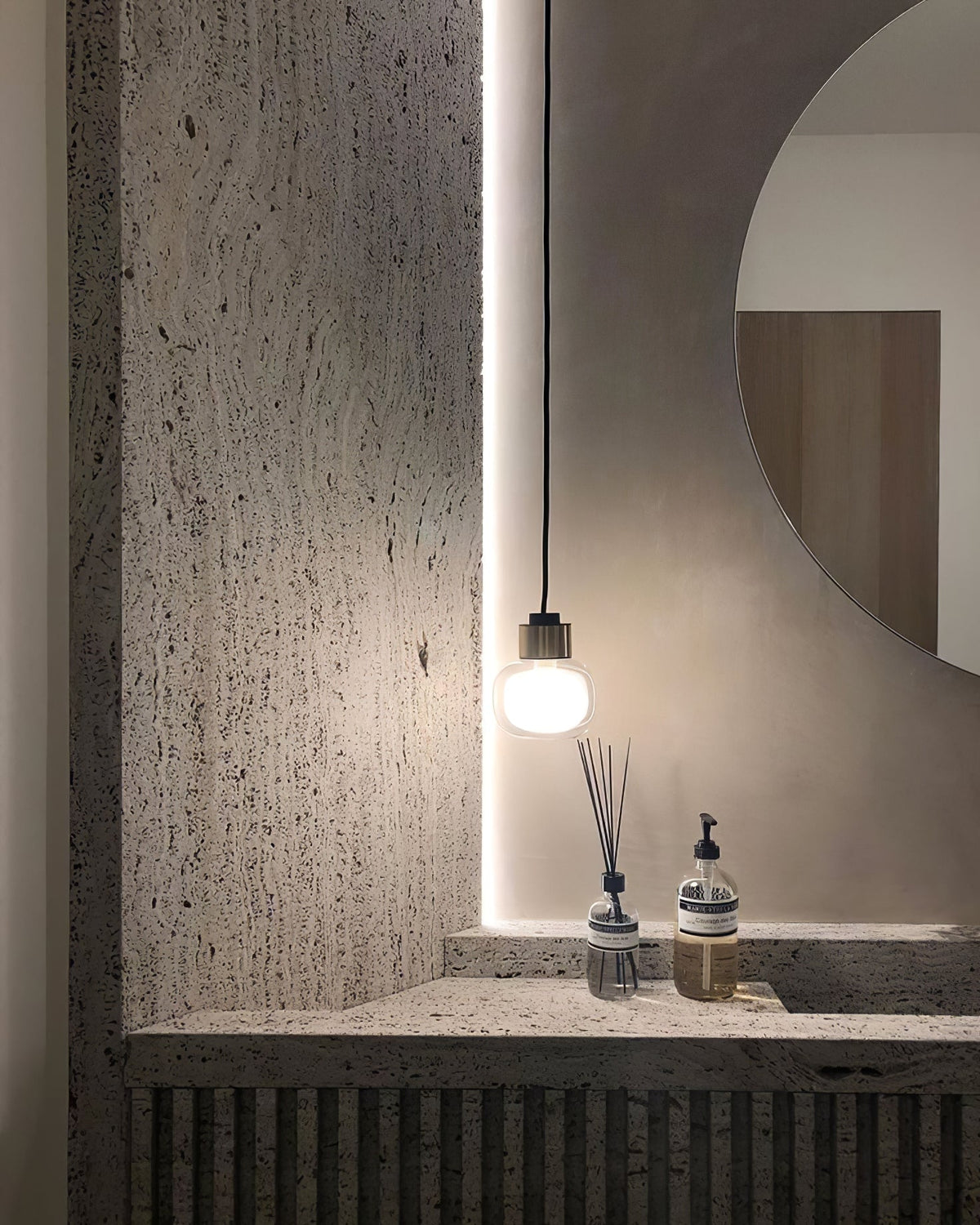 A Clérachic glass pendant glows beside a pale round mirror above a textured stone vanity with a diffuser and a soap bottle.