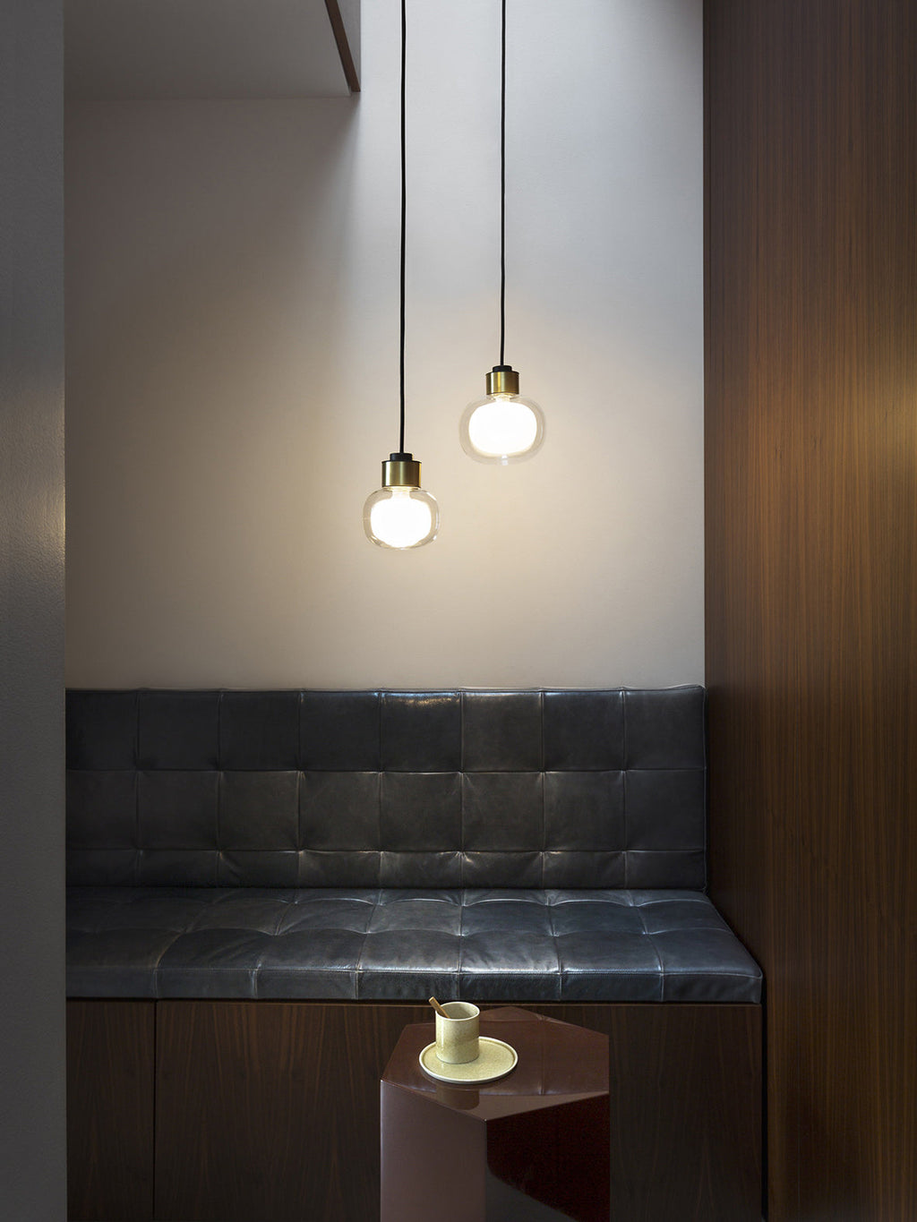 Two Clérachic glass pendants hang at different heights above a dark tufted bench, beside a walnut panel and a small side table.
