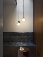 Two Clérachic glass pendants hang at different heights above a dark tufted bench, beside a walnut panel and a small side table.
