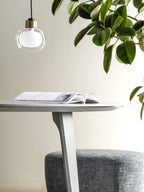 A Clérachic glass pendant hangs beside a large green plant above a pale round table with an open book and a grey pouffe.
