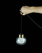 A hand lifts the cord of a Clérachic glass pendant against a black background, showing its clear glass globe and brass collar.