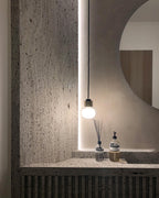 A Clérachic glass pendant glows beside a round mirror over a textured travertine-style stone shelf, with a diffuser and a soap pump.