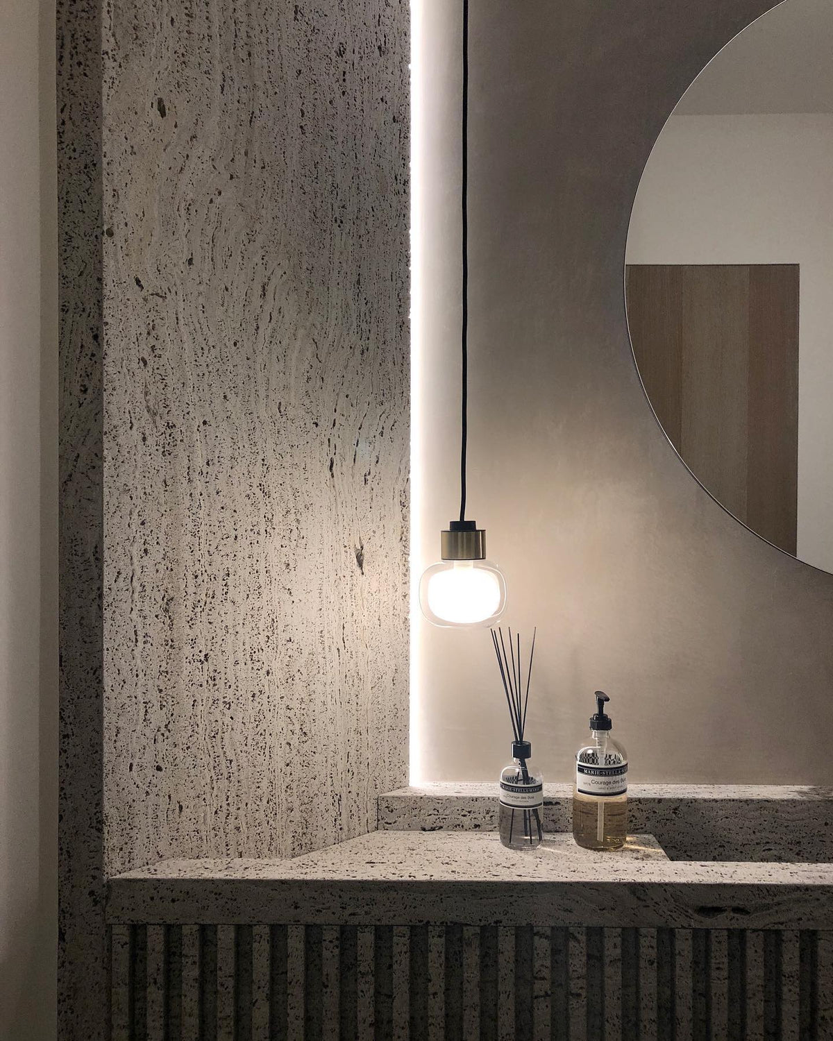 A Clérachic glass pendant glows beside a round mirror over a textured travertine-style stone shelf, with a diffuser and a soap pump.