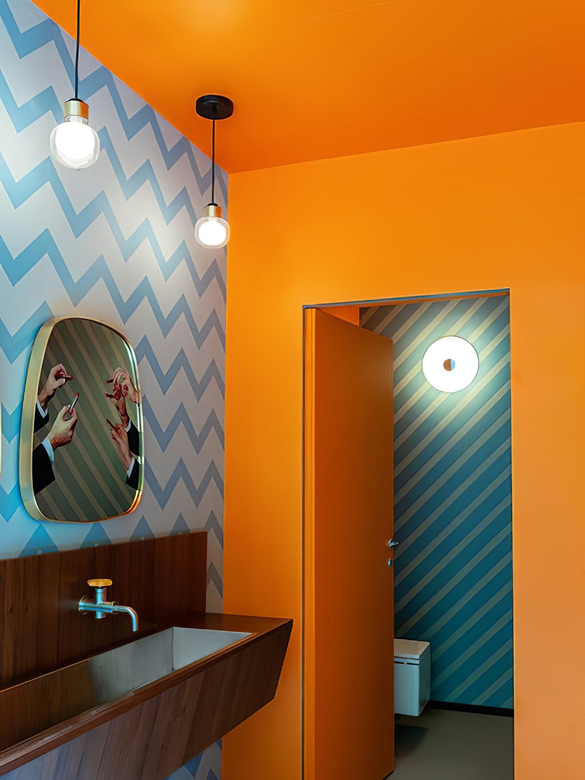 Two Clérachic glass pendants hang in a bathroom with bright orange walls, blue chevron wallpaper and a wooden basin.