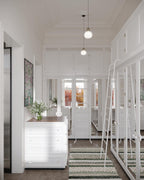 Two Clérachic glass pendants hang in a bright hallway lined with white panelling and a white chest of drawers, a striped rug below.