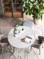 A Clérachic glass pendant hangs over a round pale-grey table with a magazine, seen from above, near a big plant and a display cabinet.