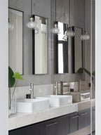 Several glass pendants from Clérachic hang in front of tall mirrors, above two white basins on a marble-topped vanity.