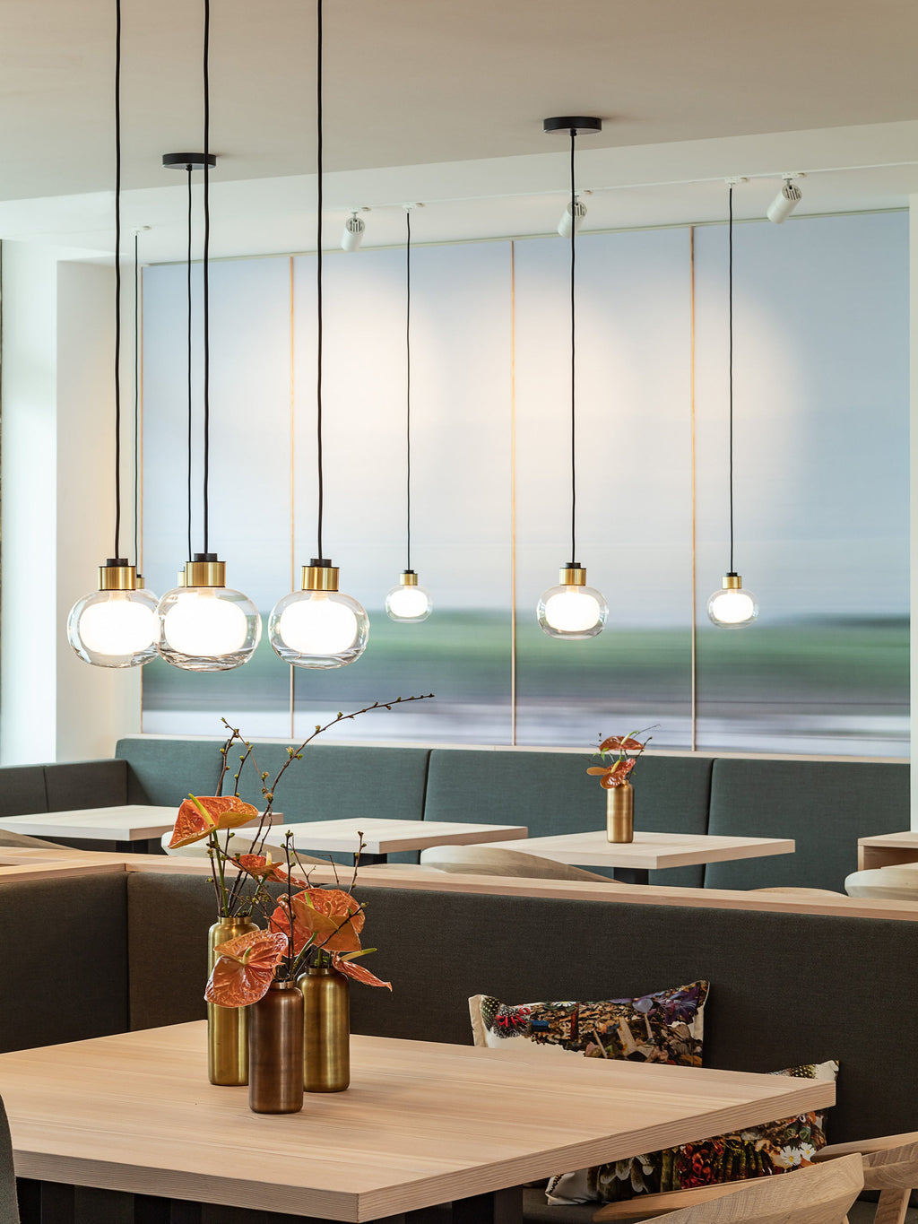 Three Clérachic glass pendants hang over a restaurant table with a vase of orange flowers, a green banquette and spotlights above.