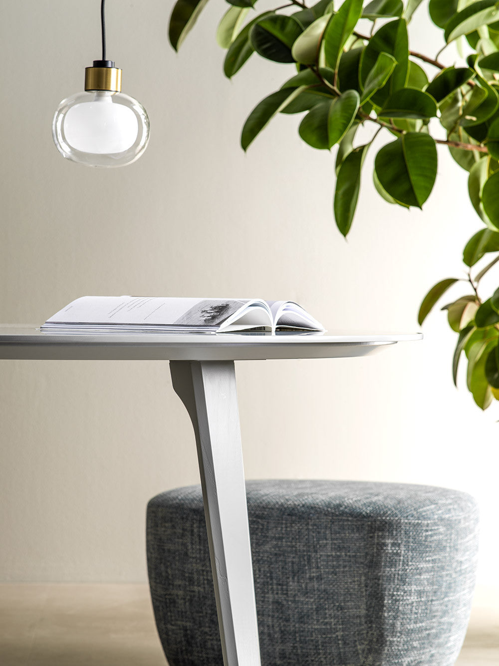 A Clérachic glass pendant hangs above a pale round table with an open book, beside a large leafy plant and a grey pouffe.