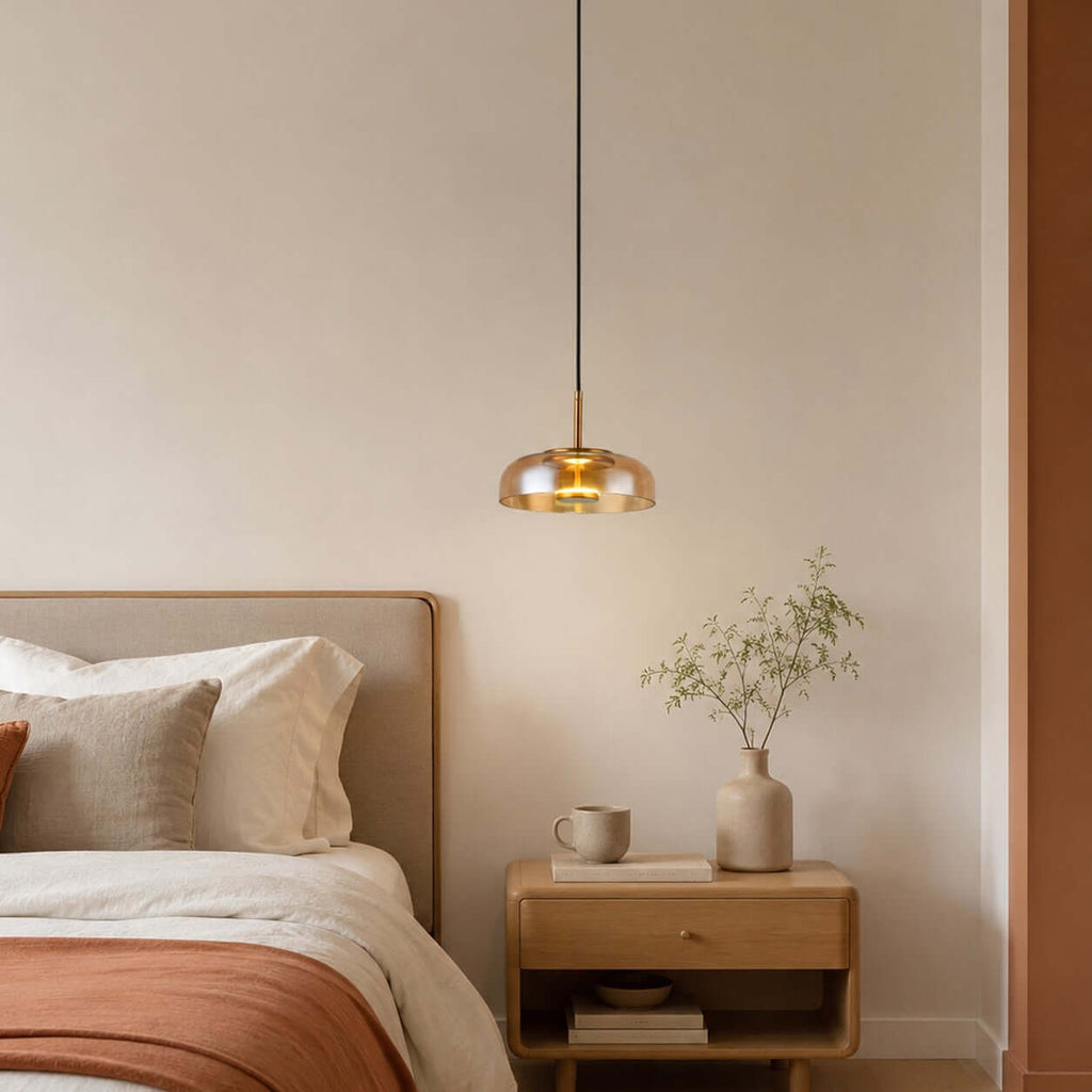 Amber glass Clérachic pendant light hanging above a bed with a terracotta throw, a wooden bedside table and a vase of twigs.