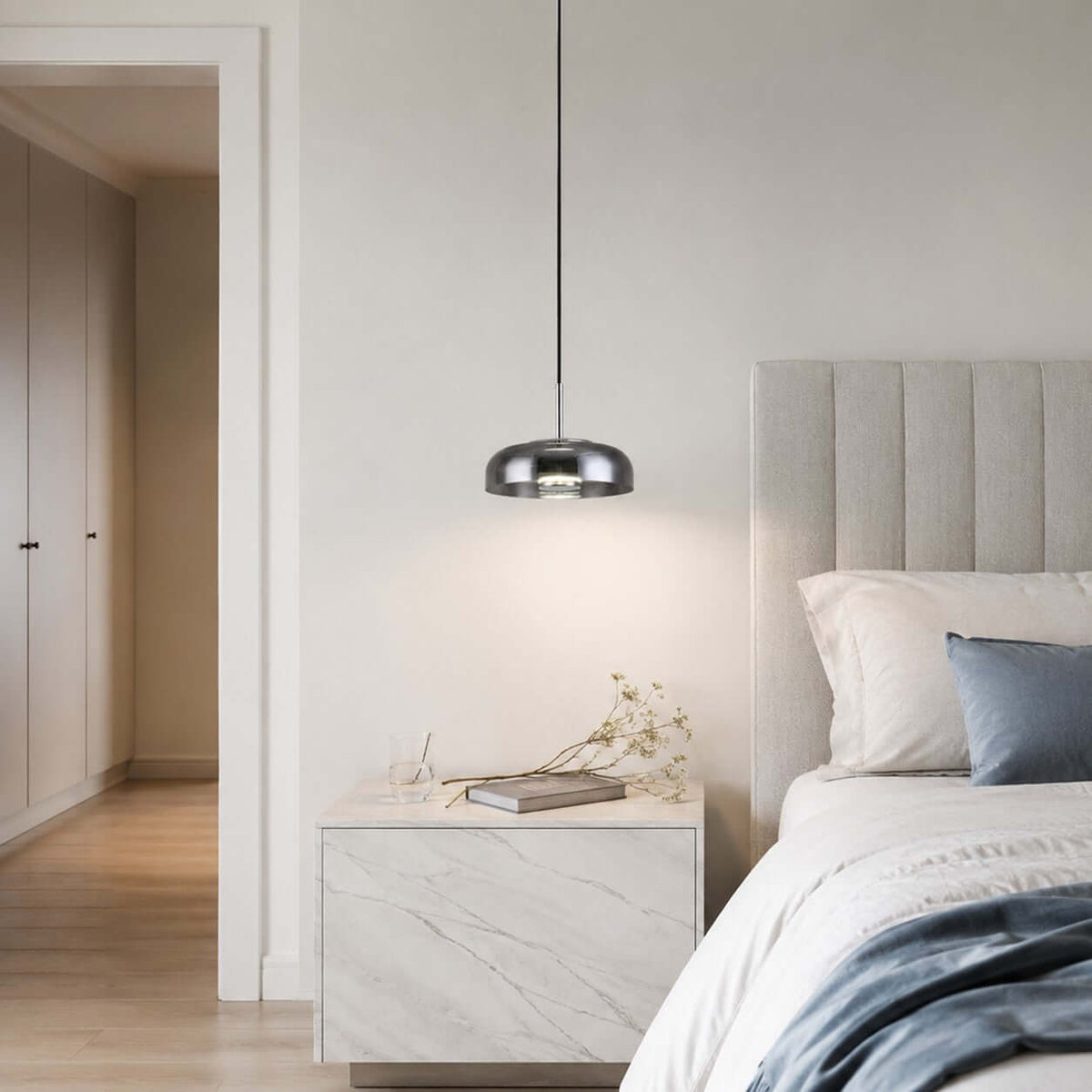 Smoke grey glass Clérachic pendant light hanging over a marble bedside table, with a ribbed headboard and blue bedding beside it.