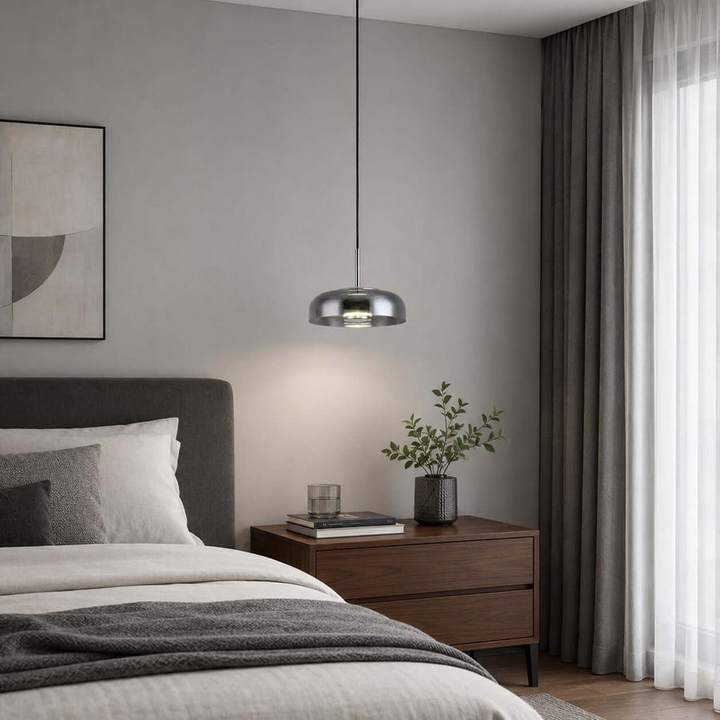 Smoke grey glass Clérachic pendant light in a grey bedroom, beside long curtains, a dark wood bedside table and a green plant.