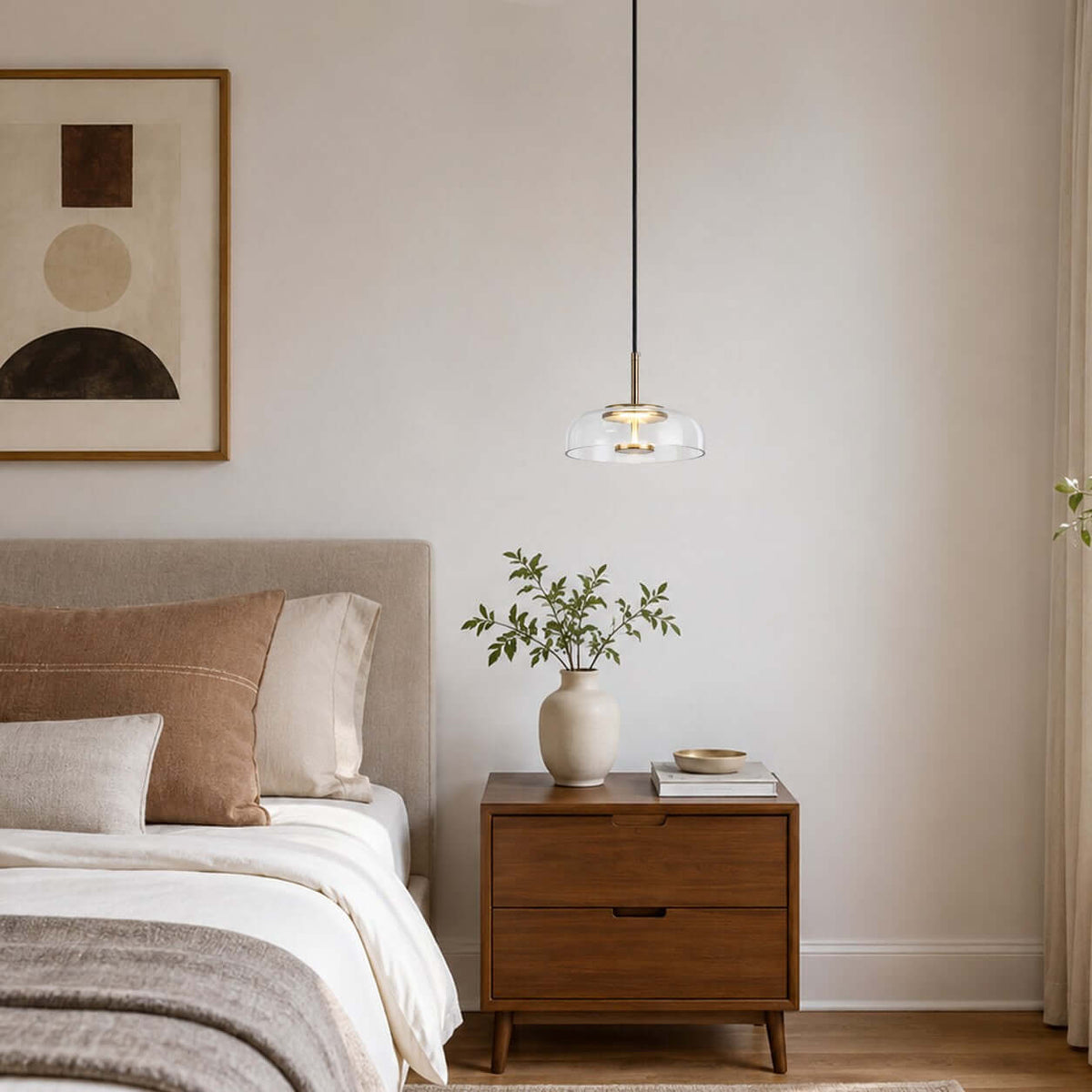 Clear glass Clérachic pendant light above a bedside table with a leafy stem in a vase, and an abstract print on the wall.