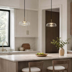 Two clear glass Clérachic pendant lights above a marble kitchen island, with white cabinets and a bowl of green apples below.