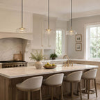 Three clear glass Clérachic pendant lights in a row above a marble kitchen island with four upholstered bar stools.